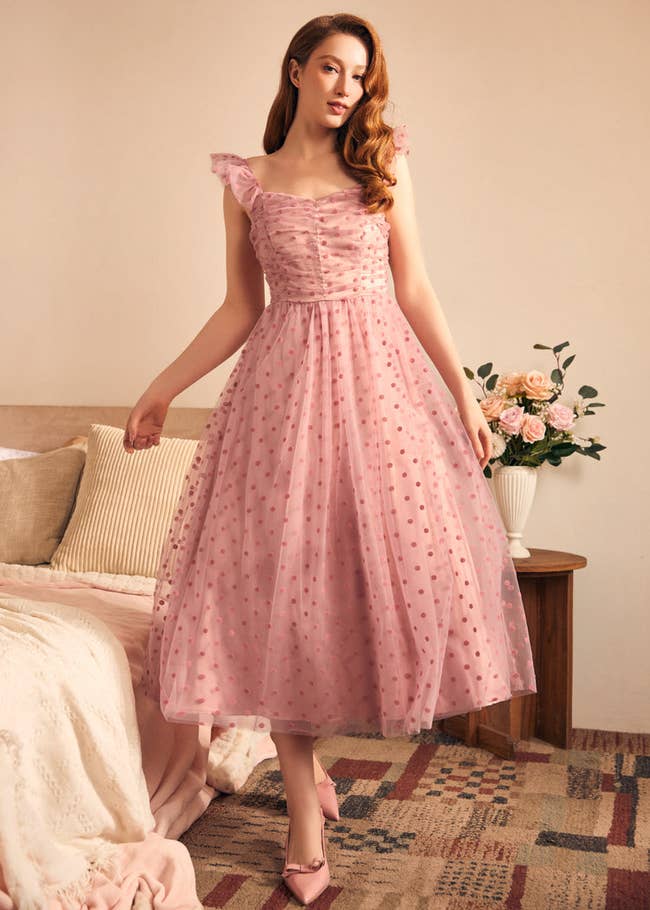 Person in a pink polka dot dress stands in a bedroom near a bed and a vase of flowers