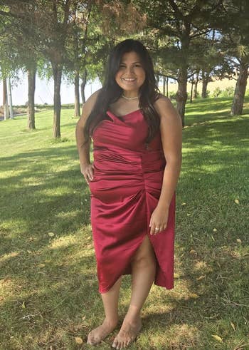 Person outdoors in a sleeveless, elegant satin dress with a draped design, posing in a grassy area with trees in the background