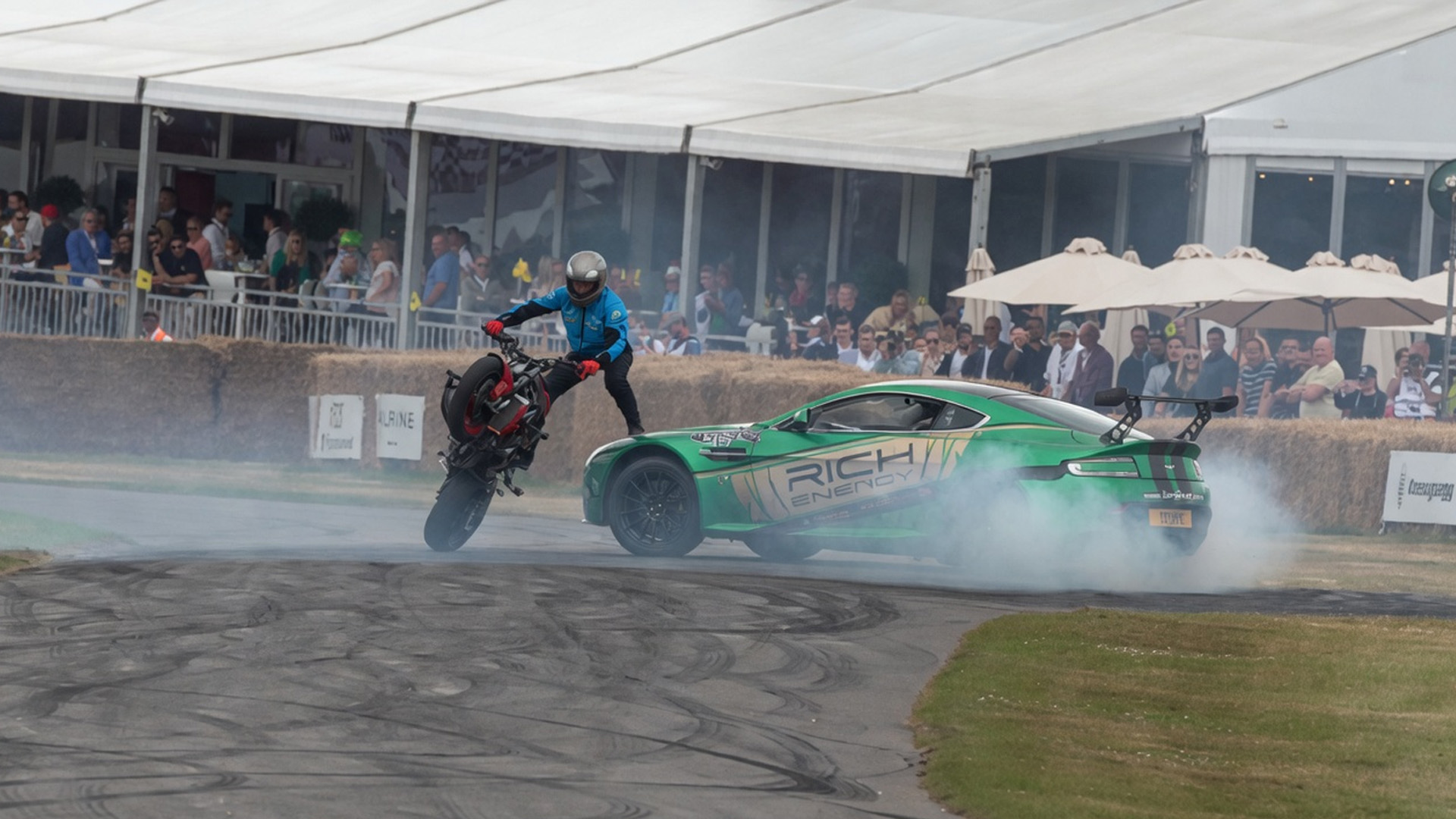 Stunt rider wheelie next to drifting Aston Martin