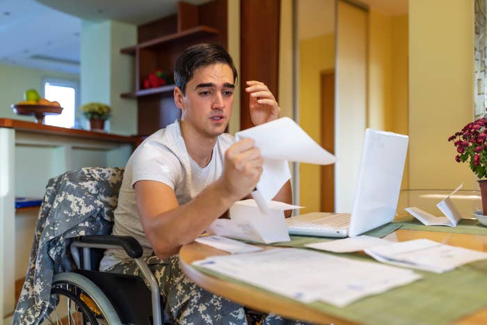 Person in a wheelchair reviewing papers at a table with a laptop, appearing concerned, in a home setting