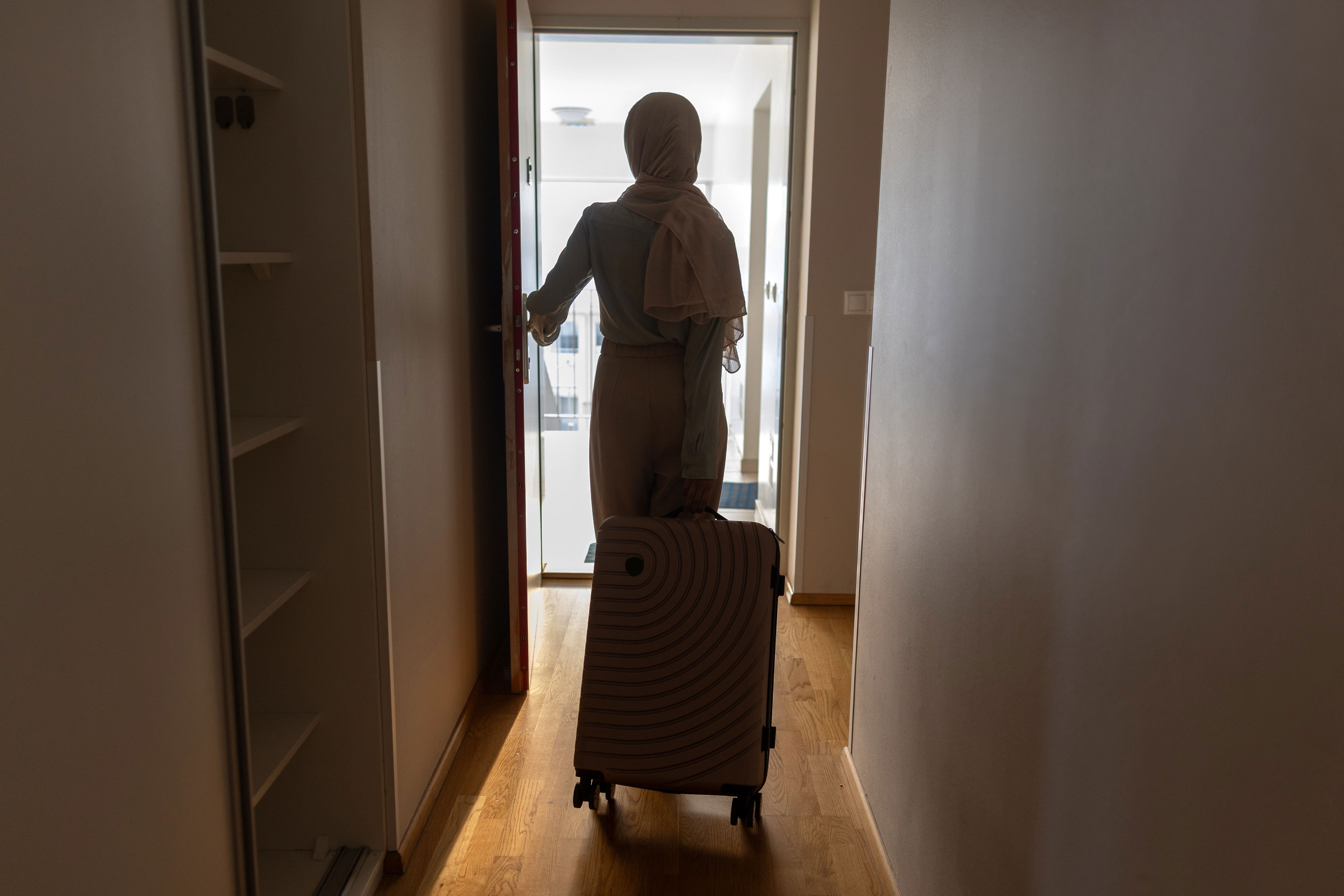 Person with suitcase exits room, heading towards bright hallway, suggesting travel or business trip