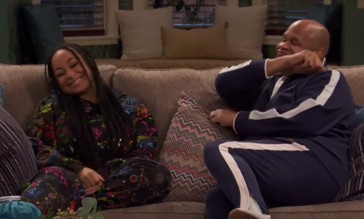 Two people sit on a couch, smiling and casually dressed. One wears a patterned outfit, the other a tracksuit. Cozy living room setting