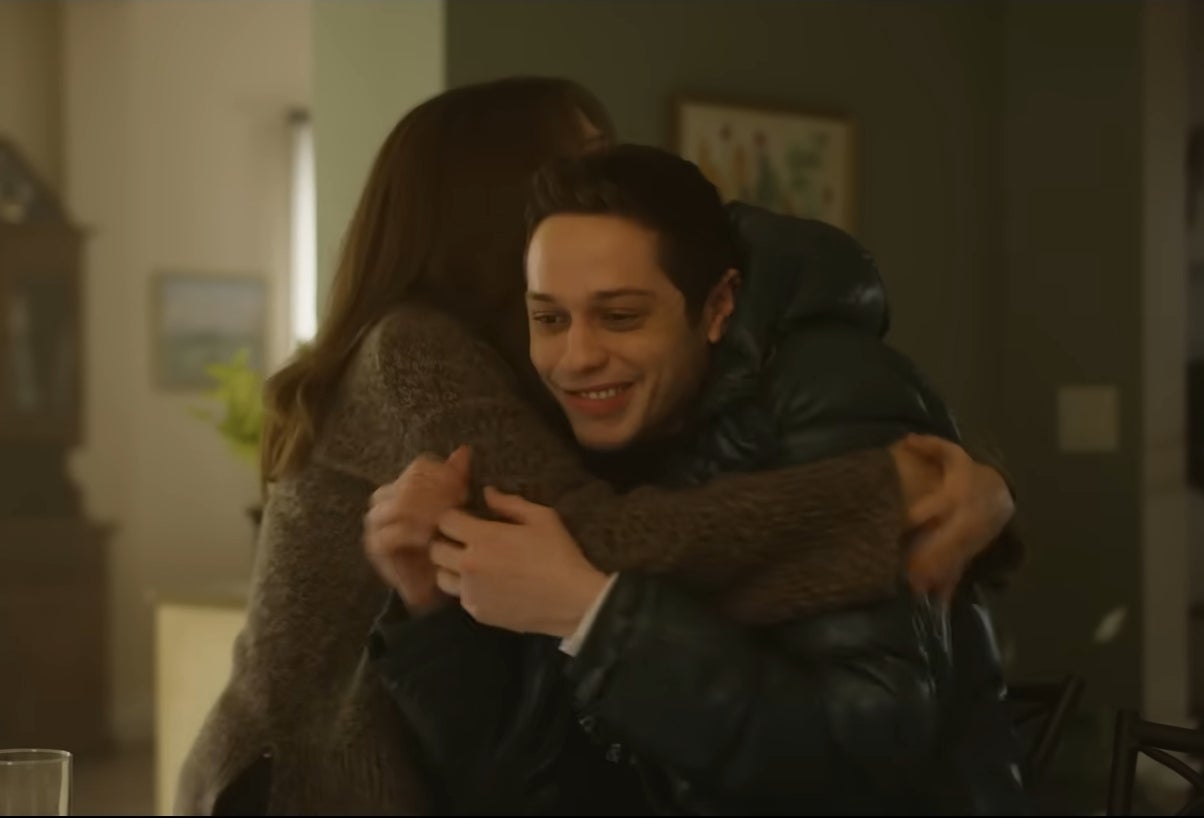 Two people hugging joyfully in a cozy room, one wearing a padded jacket. A wall clock is visible in the background. Appears to be a scene from "Bupkis."