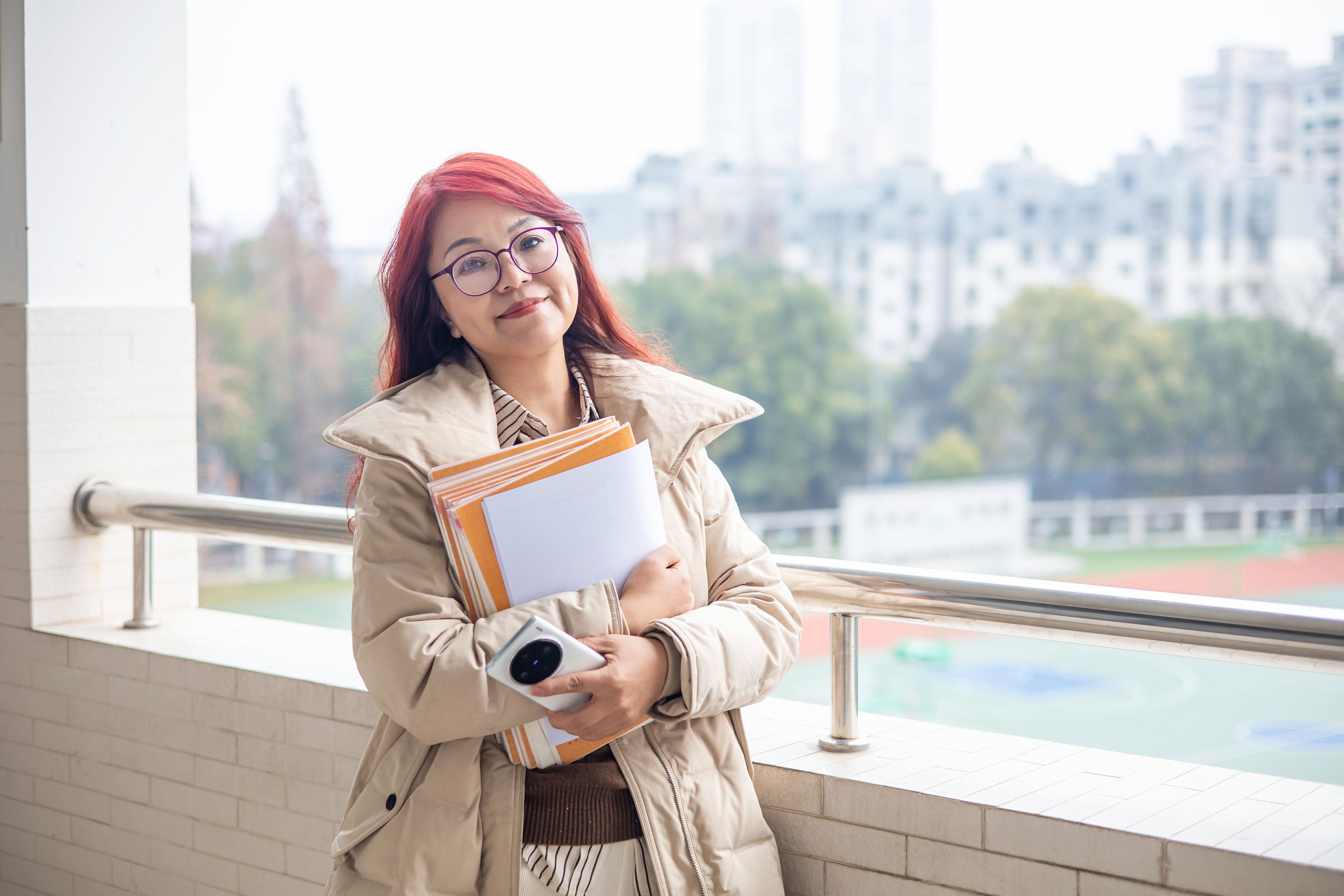 Person smiling, holding folders and papers, standing on a balcony overlooking a cityscape