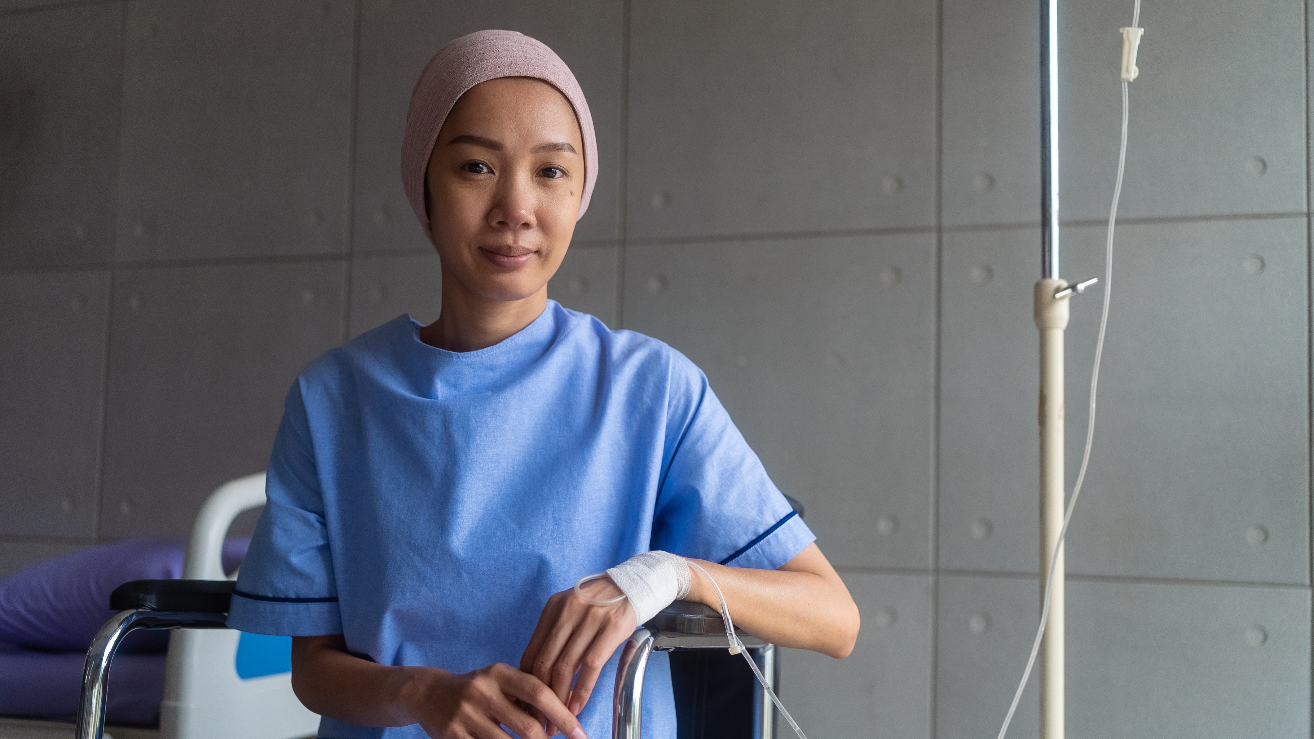 Person in a hospital room wearing a medical gown, seated with an IV stand and bandaged hand, conveying recovery or healthcare context