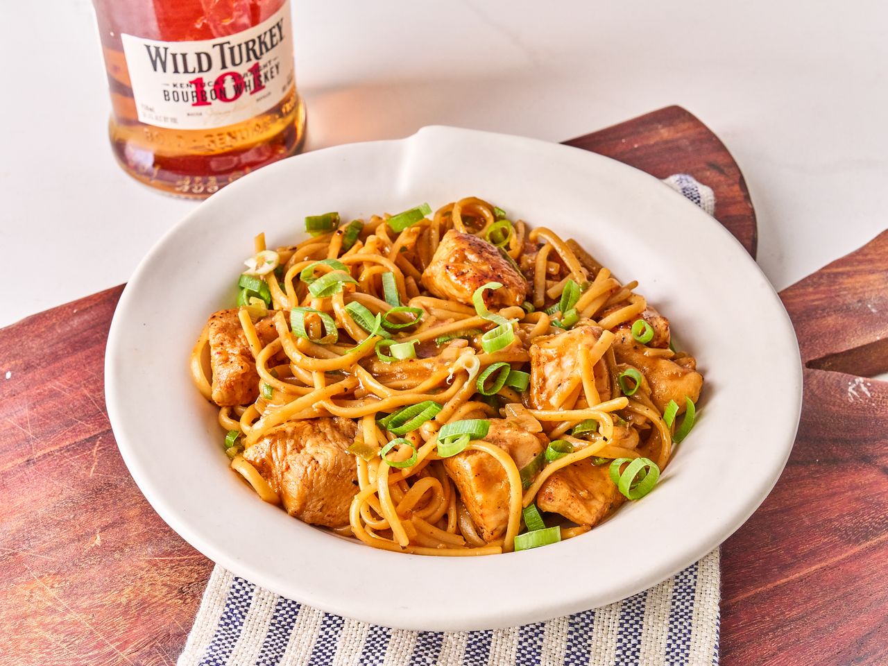 Bowl of chicken and noodles garnished with green onions on a wooden surface, bottle of whiskey in the background