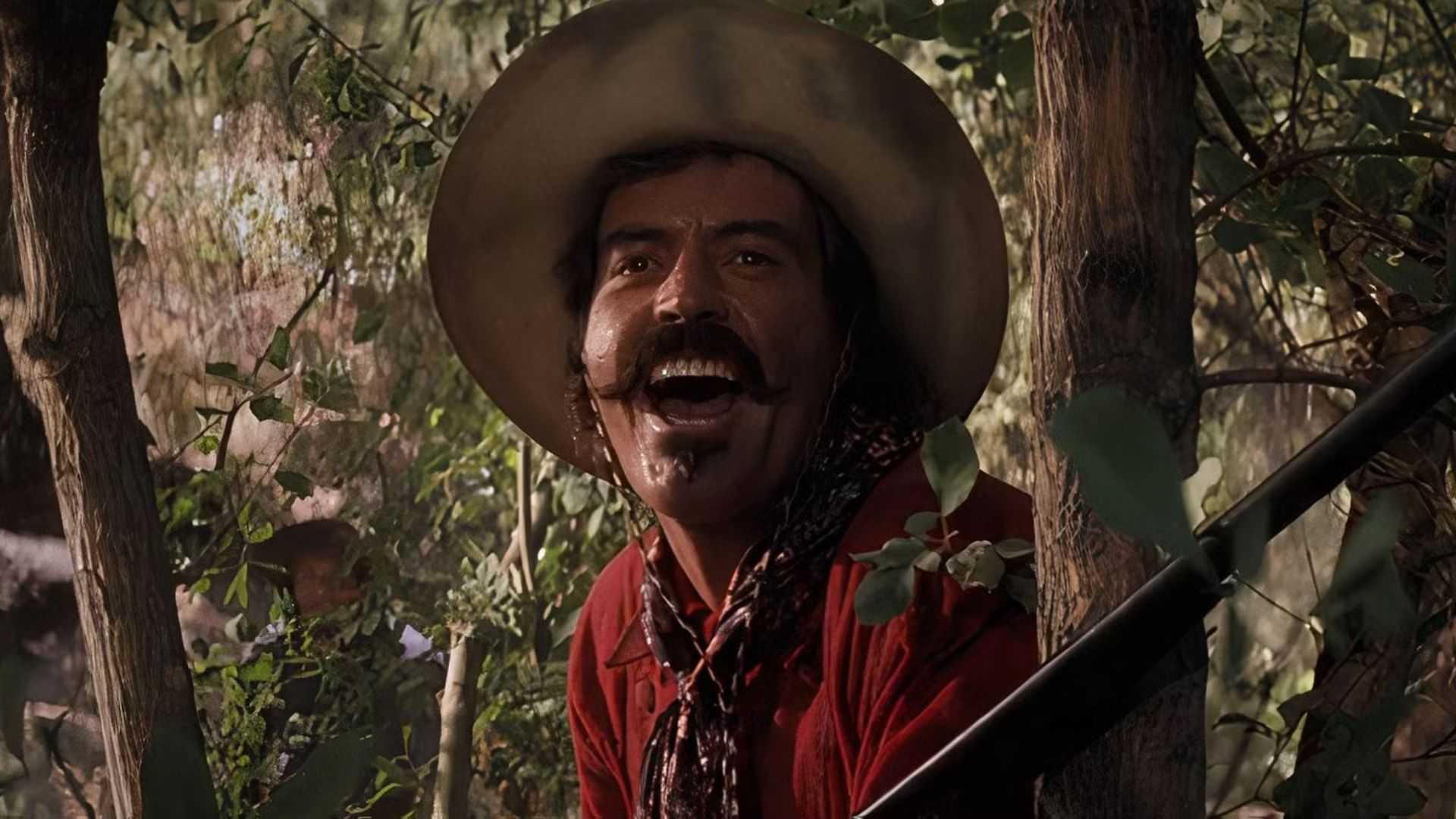 Curly Bill Brocius (Powers Boothe) cackling while hiding behind a tree and holding a shotgun in Tombstone