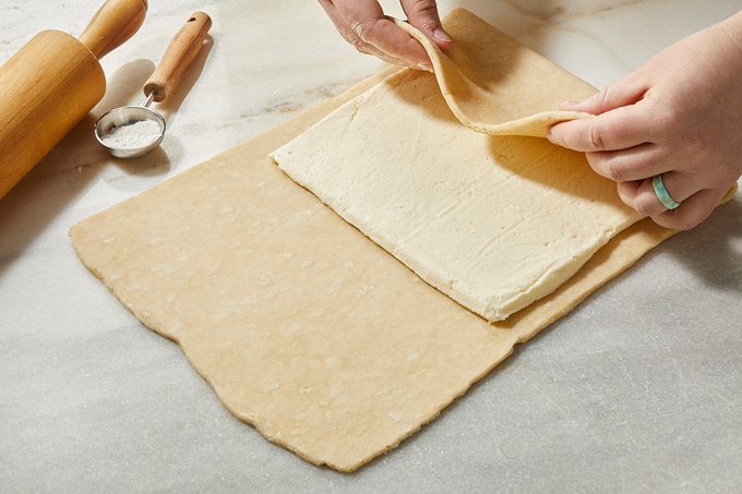 Hands folding a sheet of dough over a square of butter on a marble surface, with a rolling pin and small dish nearby, preparing laminated dough for baking.