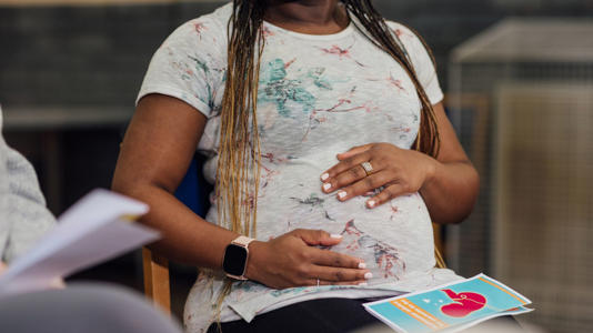 A medium close up over the shoulder view of a blooming 27 weeks pregnant woman holding her pregnancy tummy in her hands as she sits in a. an