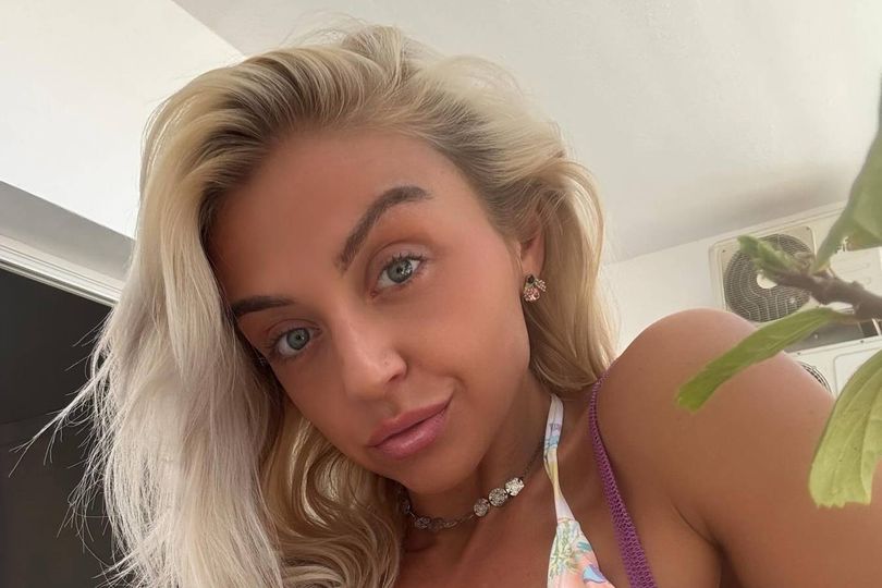 An individual with light blonde hair is captured in a close-up, wearing a sleeveless top of a vibrant purple hue, adorned with a floral pattern. The setting appears to be indoors, with a blurred background suggesting a leafy plant. The individual is looking directly at the camera, exuding a sense of confidence.