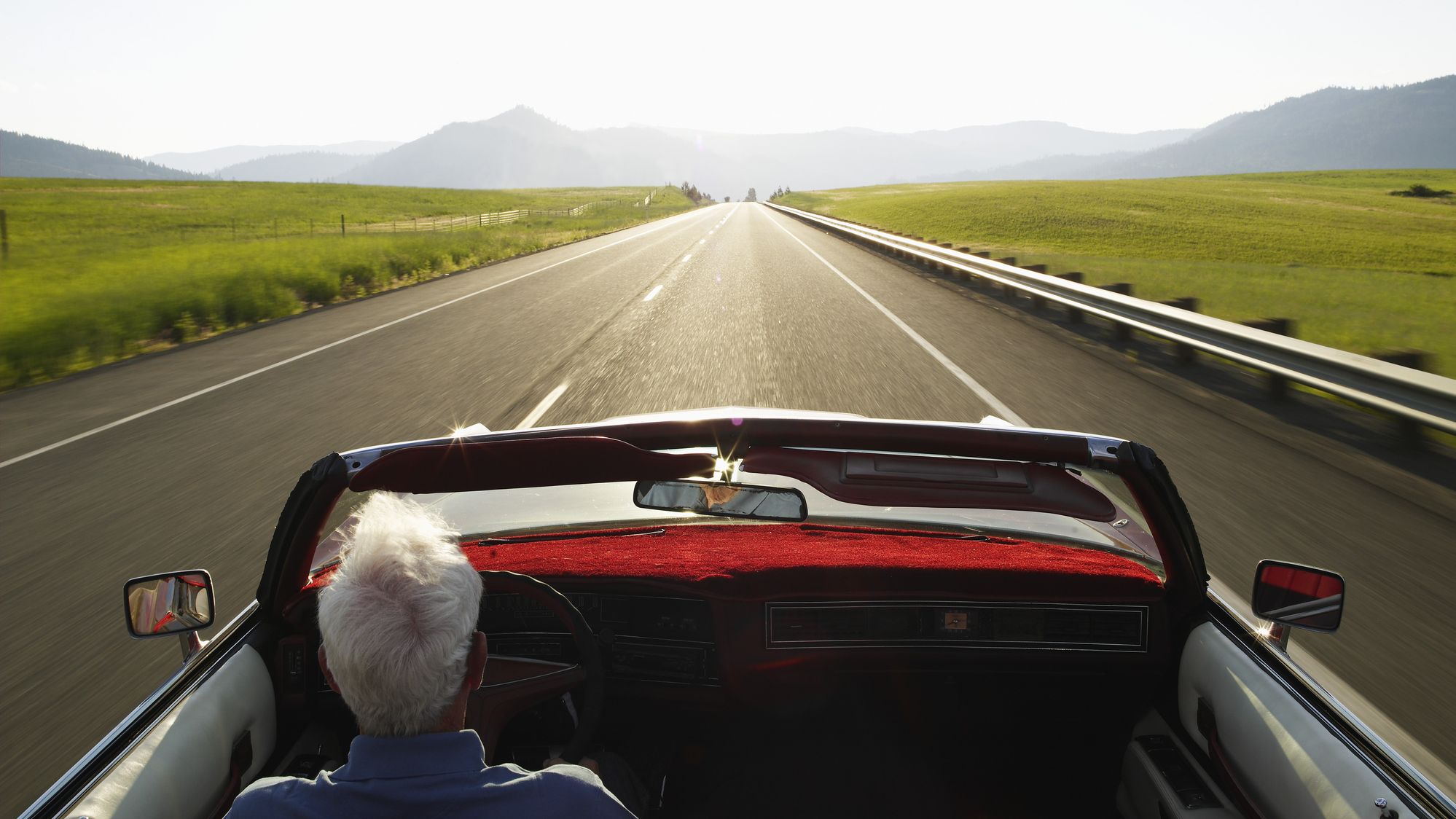 A senior man drives a convertible car down a long, open road.