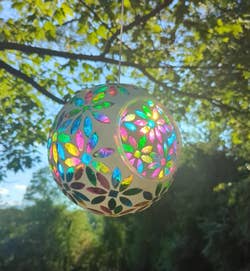 Decorative, spherical hanging ornament with a floral cutout design, featuring multicolored reflective elements, set against a natural outdoor backdrop