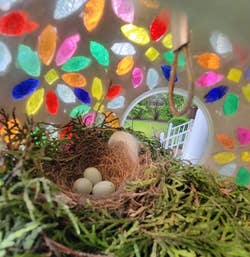 Bird's nest with three eggs inside a lantern adorned with colorful floral patterns; a peaceful garden is visible outside