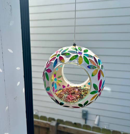 Hanging spherical bird feeder with colorful leaf-shaped designs, filled with multicolored seeds, against a fence backdrop