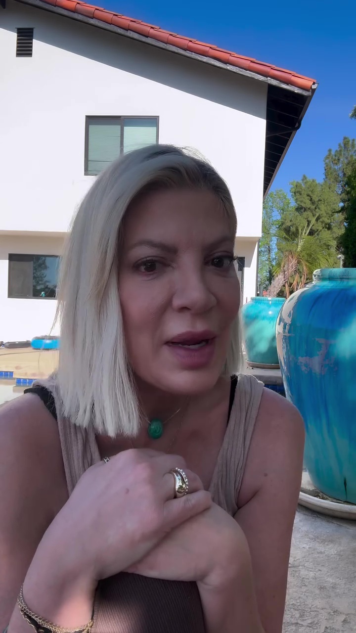 Tori Spelling speaking outdoors in front of a white building and blue planters.