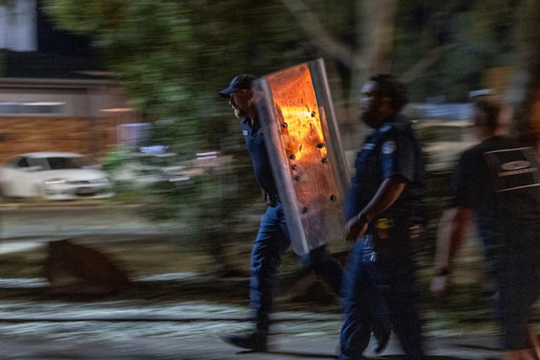 A police officer carries a large riot shield.