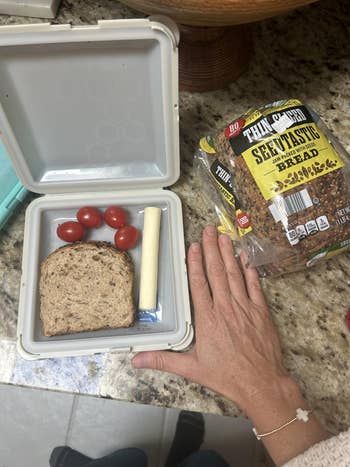 Sandwich lunchbox with tomatoes and cheese stick beside a hand and seeded bread bag on the counter