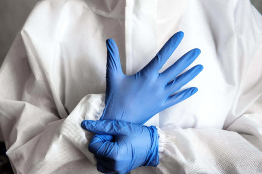 Doctor or nurse in white medical PPE suit puts on protective gloves, physician in hospital close-up. Concept of professional clinical care, uniform an