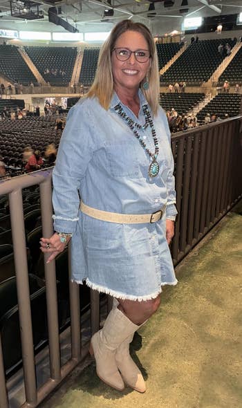 Person in denim dress with ruffled hem and knee-high boots stands by railing in an event arena