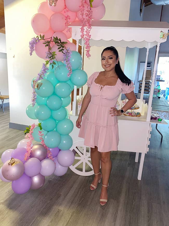 Person in a pink dress stands by a decorated cart with balloons and flowers, appearing at an event