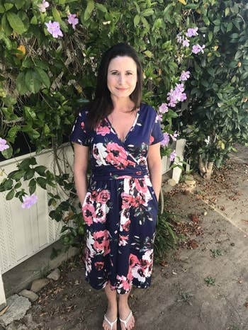 A reviewer wearing a floral dress stands outside in front of a leafy background with purple flowers