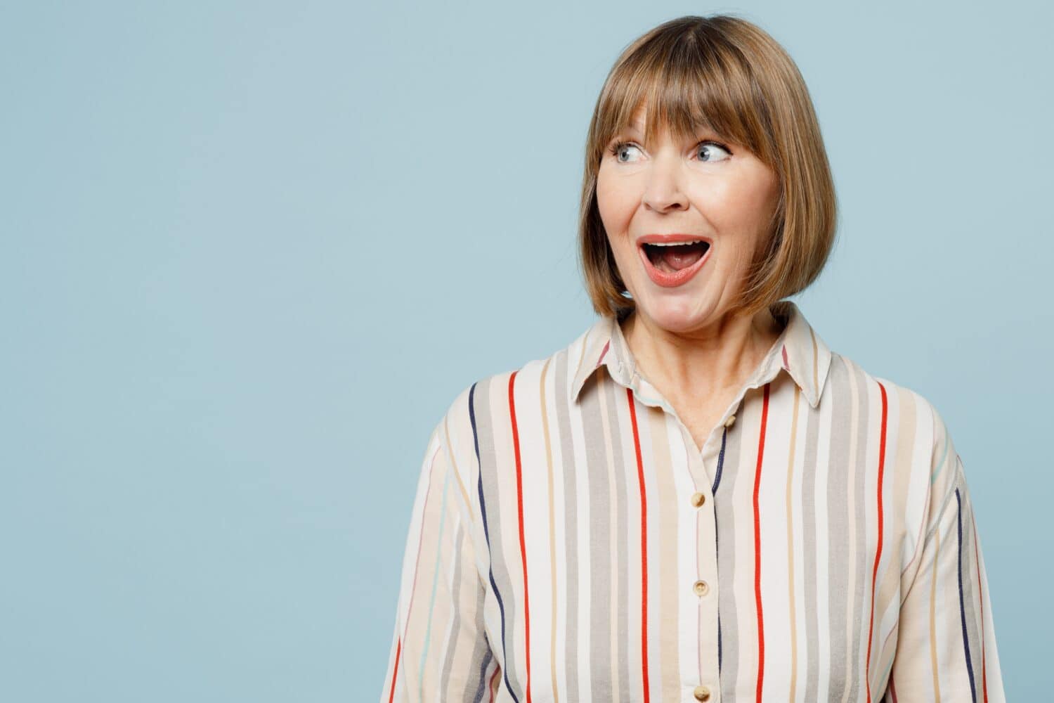Elderly shocked surprised fun woman 50s years old wear light striped shirt look aside on workspace area mock up isolated on plain pastel blue cyan color background studio portrait. Lifestyle concept