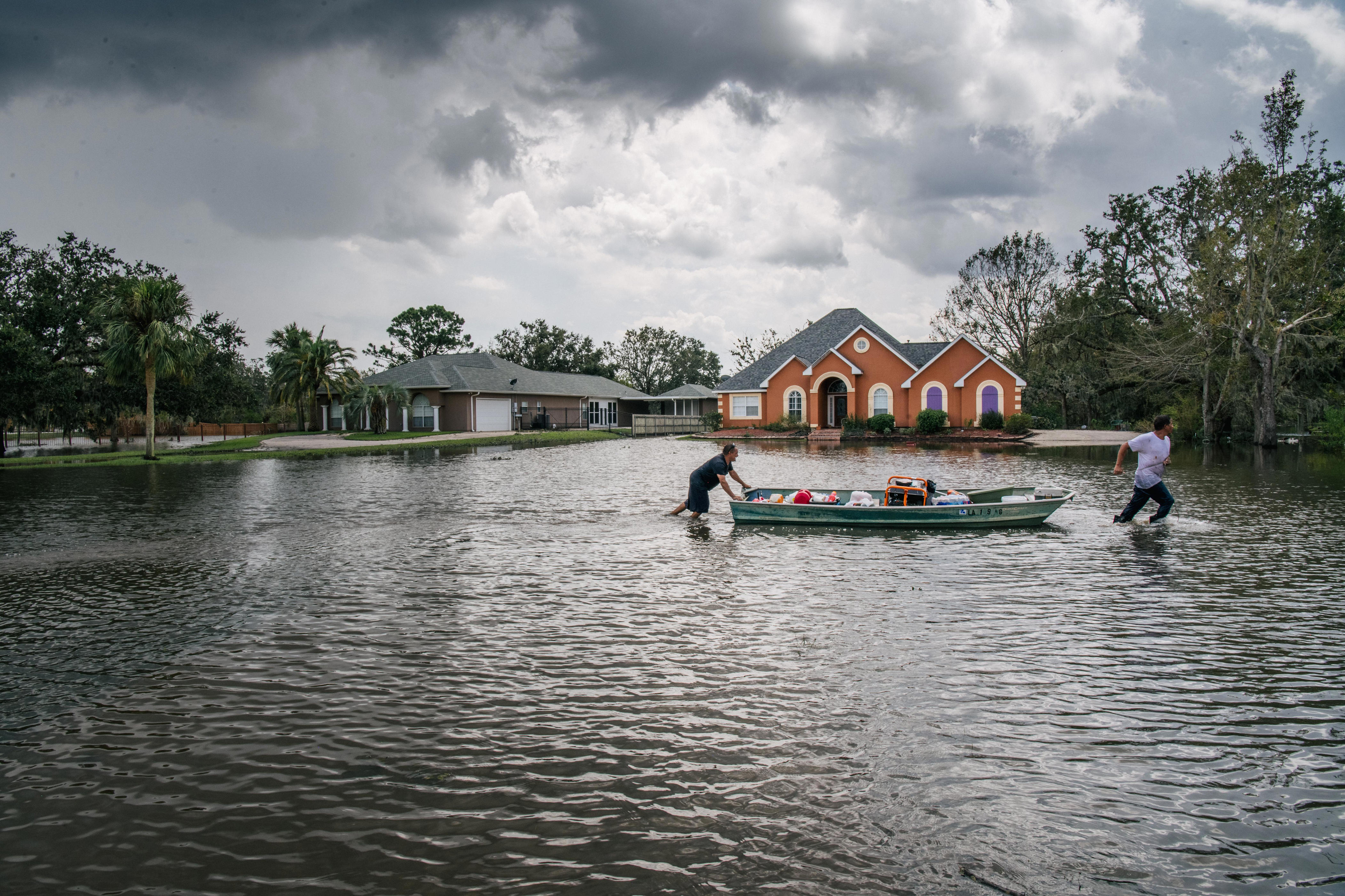 People move a boat in high flood waters with houses in the background and trees surrounding.