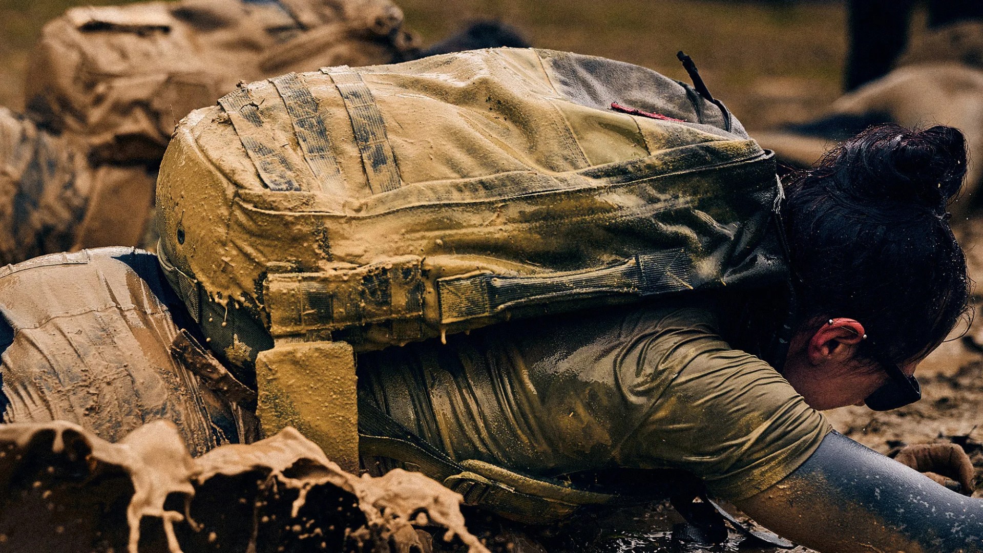 Person crawling through muddy terrain wearing a mud-covered tactical backpack and green shirt.
