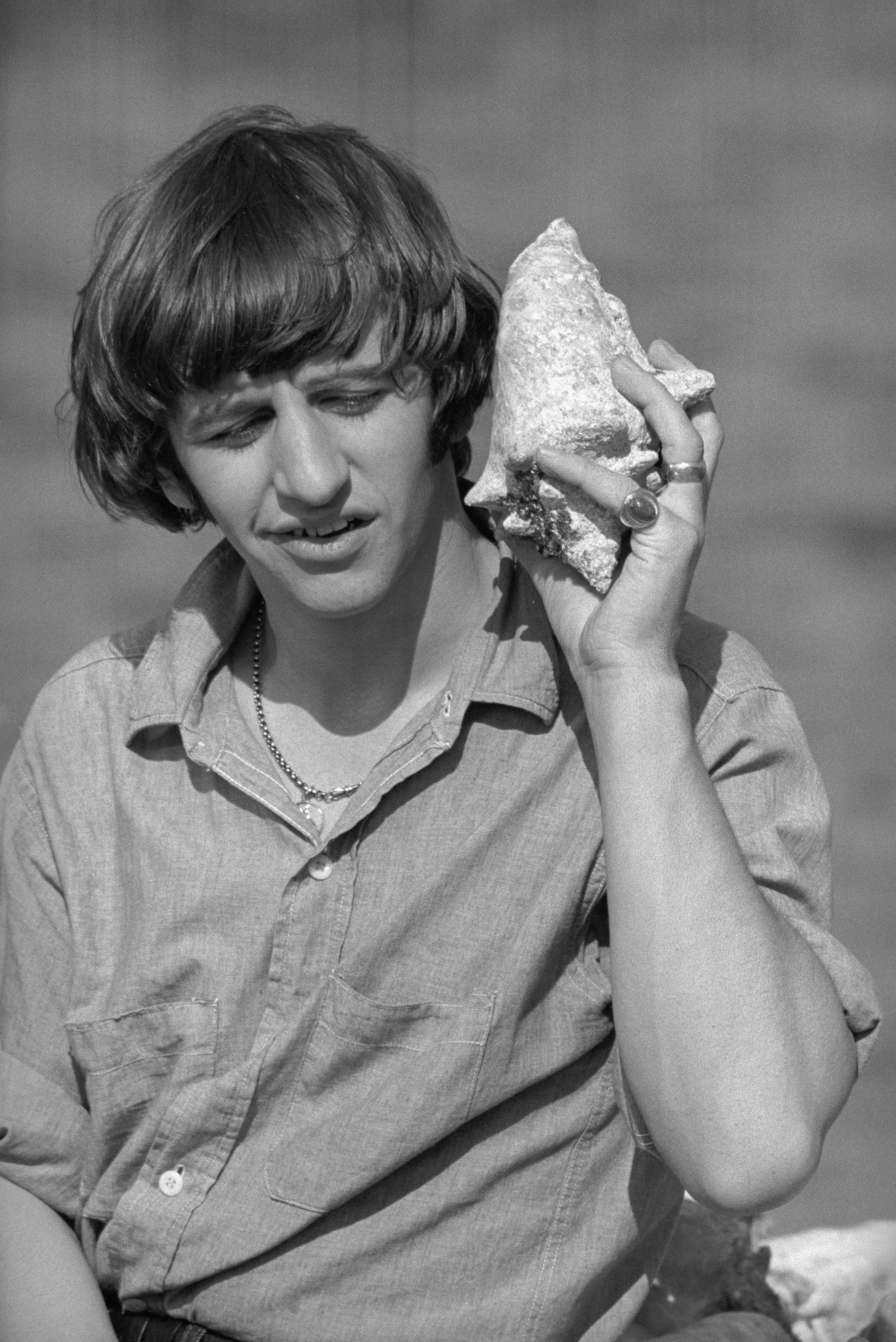 (Original Caption) A Corporation at Work. Nassau: Ringo Starr, a member of that swingin's quartet, The Beatles, listens to the chords of a sea shell during a brief break in the filming of a new movie in Nassau. Ringo, who recently gave up his bachelorhood, picked up the Conch shell while doing some scenes at the Bayshore Marina.