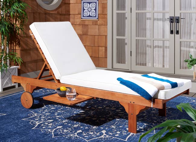 Wooden outdoor lounge chair with a white cushion, set on a patio. A towel, a glass of water, and a bowl with a lemon are nearby