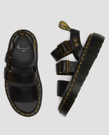 Pair of Dr. Martens black strap sandals with chunky soles and yellow stitching, laid out for a shopping feature