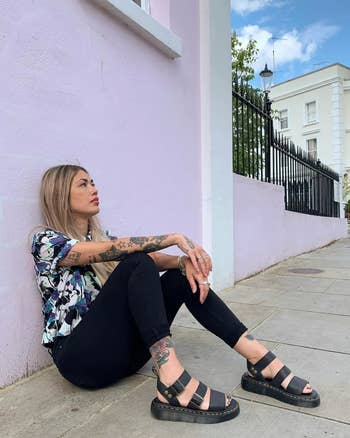 Person sitting on sidewalk, wearing strappy sandals, black pants, and a patterned shirt, showcasing casual street style