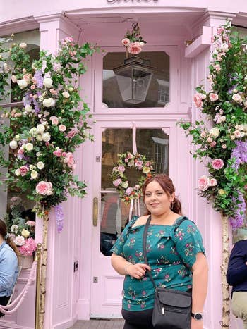 Reviewer's photo of themself standing in front of a flower-adorned pink doorway, wearing a floral top and carrying the bag in the color Black