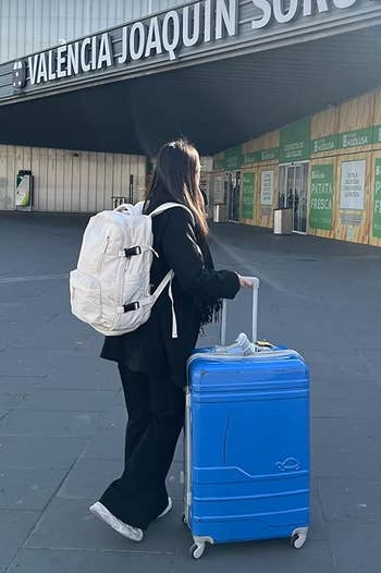 Another reviewer's photo of them wearing the backpack in the color Beige and pushing a blue rollerboard suitcase outside of a train station