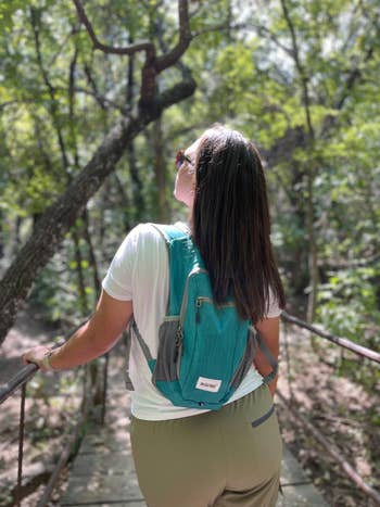 Person with a small backpack stands on a bridge in a forest, looking up at the trees. Casual outdoor attire suggests a hiking or nature activity