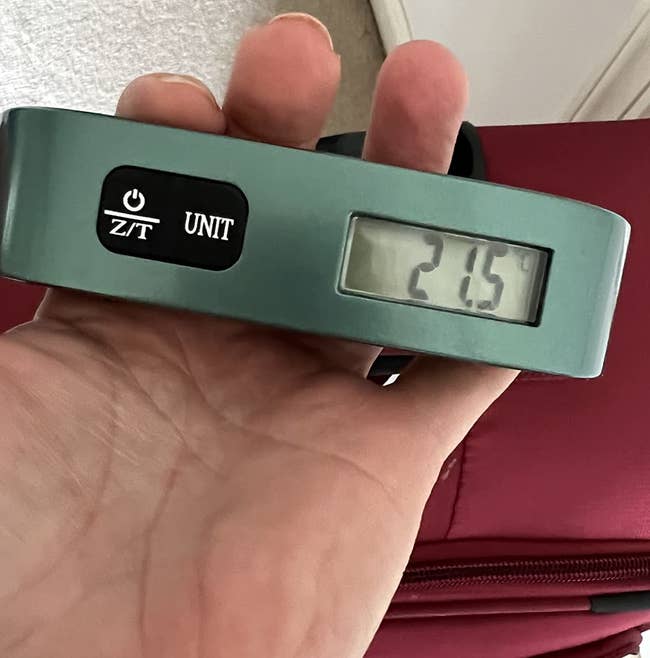 Reviewer's photo of their hand holding a digital luggage scale displaying 21.5, with a red suitcase in the background