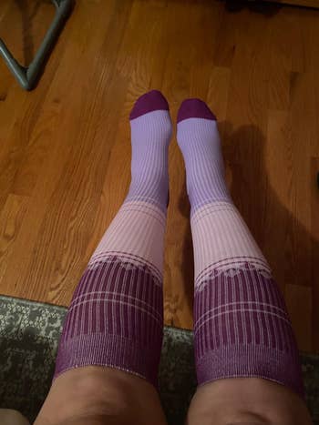 Person wearing knee-high, vertically striped compression socks with a gradient pattern, sitting on a wooden floor