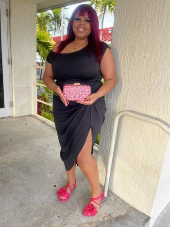 Person in a stylish black dress with a slit, holding a pink patterned clutch, and wearing matching pink sandals, standing by a wall outdoors
