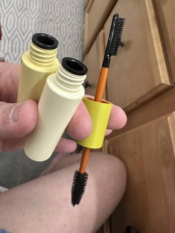 A person holds an open mascara tube, displaying the applicator brush. A drawer and patterned carpet are partially visible in the background