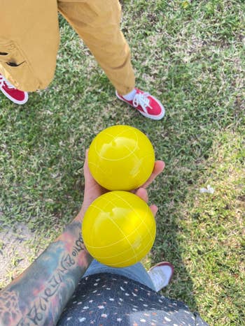 Person holding two yellow bocce balls, standing on grass near another person in casual attire and red sneakers