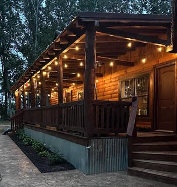 Wooden cabin with a large porch illuminated by string lights, surrounded by trees. Stone pathway leads to steps. Cozy, rustic atmosphere at dusk