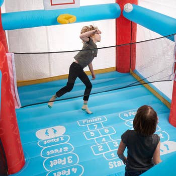 Two children play and jump in a blue inflatable basketball-themed playpen with a mini hoop and netted sides, enjoying an interactive game