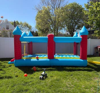 Inflatable bounce house with turrets in a backyard setting on a sunny day. A toy car and scattered balls are on the grass nearby