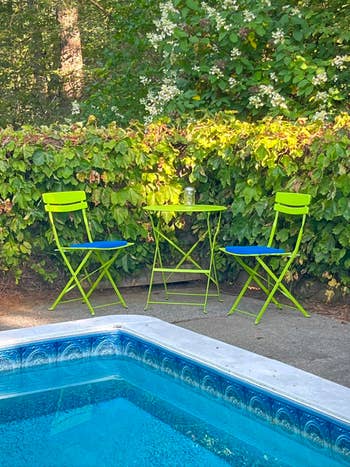 Two green folding chairs and a matching table are set by a pool, surrounded by lush greenery, suggesting a relaxing outdoor space