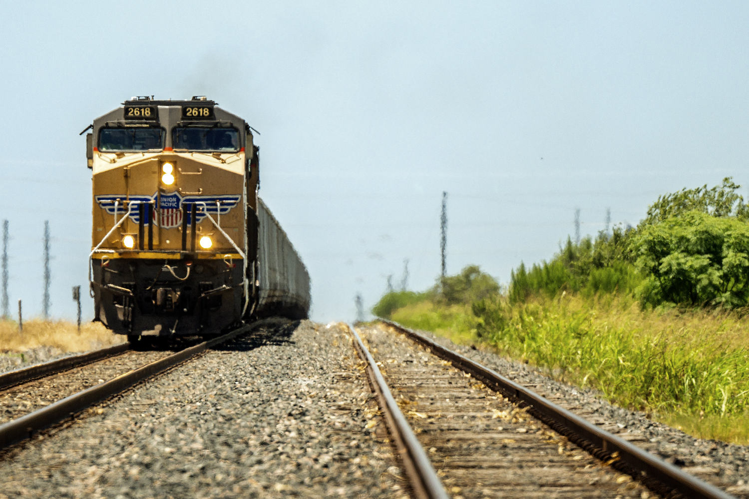 Shimmering heat distorts a view of a yellow Union Pacific freight engine approaching with its headlights on in bright sunlight.