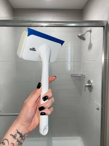 Person holds a Mr. Clean shower brush inside a modern bathroom with a glass-enclosed shower