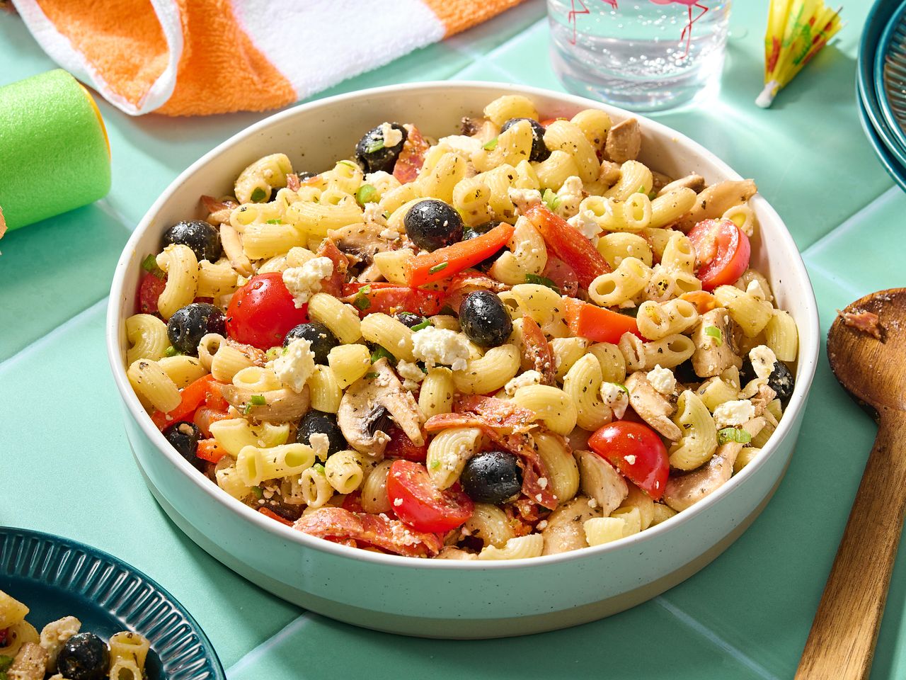 A bowl of Greek pasta salad with ingredients like tomatoes, olives, and feta cheese visible on a table setting