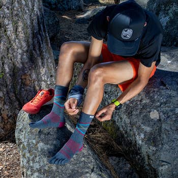 Person sitting on a rock, wearing five-toe socks, adjusting them next to a pair of trail shoes. Outdoors setting, suggesting readiness for hiking
