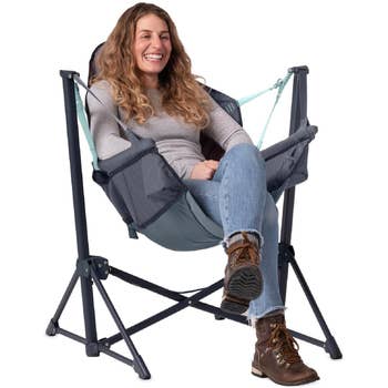 Laughing person sitting in a cozy hammock-style camping chair, wearing a gray sweater, jeans, and boots