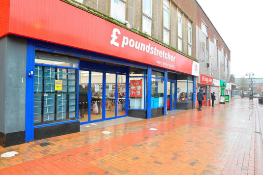 An exterior view of a commercial building with glass doors and red signage, likely a retail establishment, is situated along a street with pedestrians. The building features a combination of red and blue accents, and the sidewalk is wet, indicating recent precipitation.